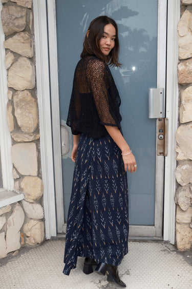 A woman standing in front of a door, wearing a long, black ikat skirt with a geometric tree pattern and a black tank top with a zero waste lace shrug, side view.