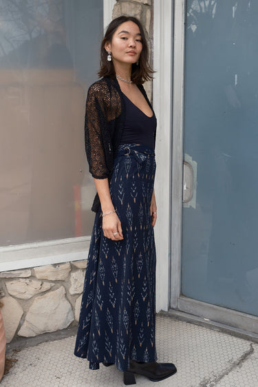 A woman standing in front of a door, wearing a long, black ikat skirt with a geometric tree pattern and a black tank top with a zero waste lace shrug, side view.