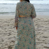 A woman standing at the beach wearing tan with green turquoise flower tapestry shirt with bell sleeves and a matching  wrap skirt. Back View