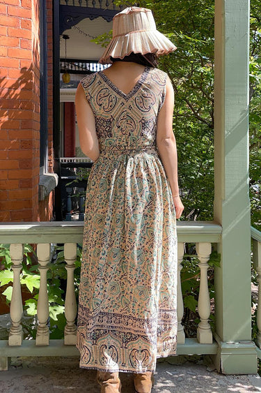 A woman standing outside wearing a beige ground turquoise flower tapestry Maxi Wrap dress, sleeveless. Back View 