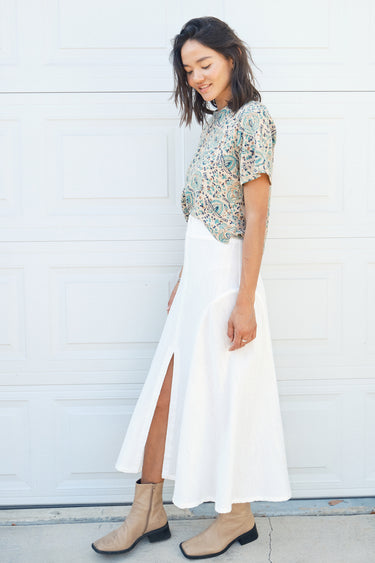A woman standing in front of a garage door, wearing a midi-length skirt in white with a floral tapestry tee top. Worn with tan ankle boots. side view