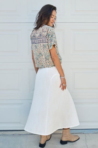 A woman standing in front of a garage door, wearing a midi-length skirt in white with a floral tapestry tee top. Worn with tan ankle boots. Back View