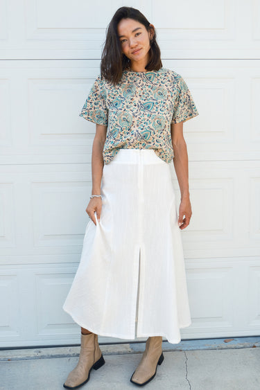 A woman standing in front of a garage door, wearing a midi-length skirt in white with a floral tapestry tee top. Worn with tan ankle boots.