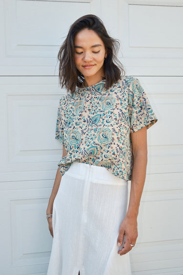 A woman standing in front of a garage door, wearing a midi-length skirt in white with a floral tapestry tee top. 
