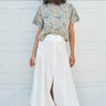 A woman standing in front of a garage door, wearing a midi-length skirt in white with a floral tapestry tee top. Worn with tan ankle boots.