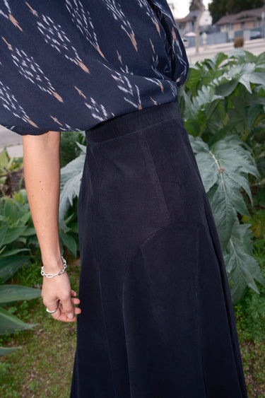 A woman standing outdoors wearing a midi black corduroy skirt with a side zipper.