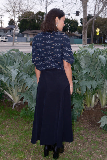 A woman standing outdoors wearing a midi black corduroy skirt with a side zipper, paired with a black top and black boots.