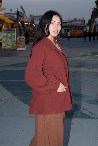 A person standing in an outdoor setting wearing a raisin unlined jacket with big patch pockets and an elastic sleeve cuff, paired with brown trousers.