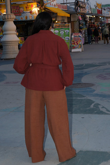 A person standing in an outdoor setting wearing a raisin unlined jacket with big patch pockets and an elastic sleeve cuff, paired with brown trousers, back view.