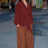 A person standing in an outdoor setting wearing a raisin unlined jacket with big patch pockets and an elastic sleeve cuff, paired with brown trousers.