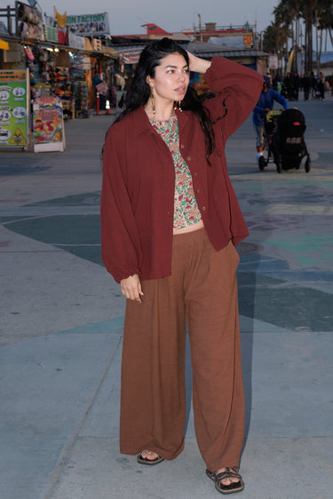 A woman standing at a beach boardwalk wearing a nutmeg brown trouser, raisin jacket and a floral print blouse. 