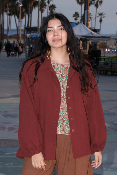 A person standing in an outdoor setting wearing a raisin unlined jacket with big patch pockets and an elastic sleeve cuff, paired with brown trousers.