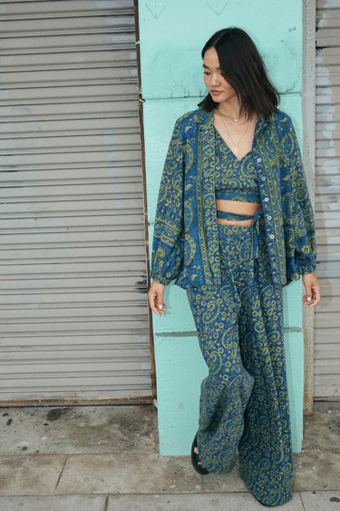 A woman wearing a blue paisley tapestry trouser with a matching Jacket and crop top, standing outside.