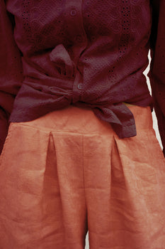 A close-up of a person wearing a dark burgundy eyelet button-up shirt tied at the waist and loose persimmon-colored pleated trousers.