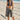 Woman in a polka dot bikini top and patterned shorts standing on a sandy beach with a pier in the background.
