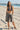 Woman in a polka dot bikini top and patterned shorts standing on a sandy beach with a pier in the background.