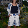 Woman standing outside wearing bermuda shorts with a white and blue tapestry print with a black tank top and a matching coat.