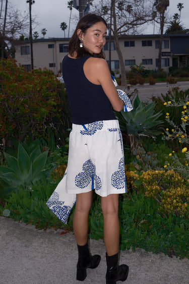 Woman standing outside wearing bermuda shorts with a white and blue tapestry print with a black tank top and a matching coat, back view.