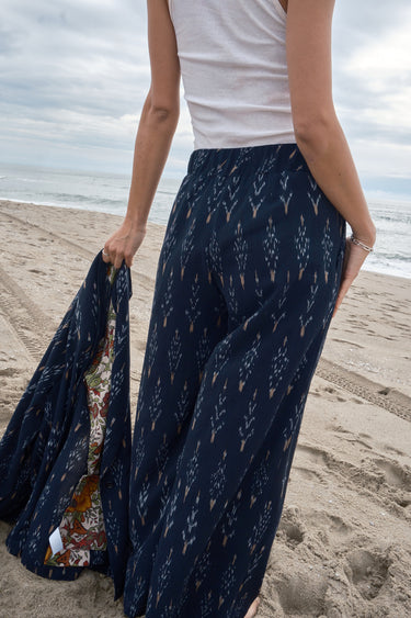 Woman at the beach wearing an ikat  low waist trousers, with white tank top and holding a matching jacket, back view.