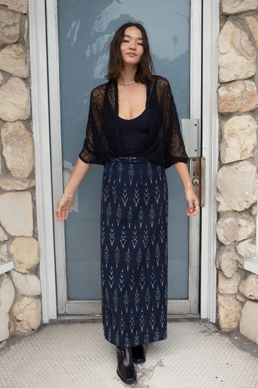 Woman standing a door way wearing a black zero waste lace shrug over a black tank top, paired with an ikat maxi skirt.