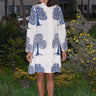 A woman standing outdoors wearing a knee-length white coat dress with blue tree patterns, featuring a hidden front button placket and pockets.
