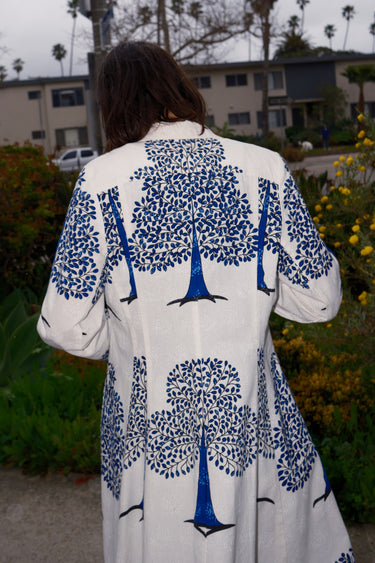 A woman standing outdoors wearing a knee-length white coat dress with blue tree patterns, back view detail.