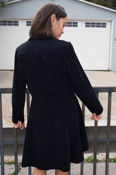 Woman standing outside wearing a black plush corduroy coat dress, back view.