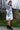 A woman wearing a white long-sleeve coat dress with a blue tree pattern, standing outdoors in front of green plants and yellow flowers.