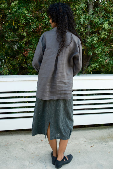 Person wearing a long gray garment standing outdoors with greenery in the background