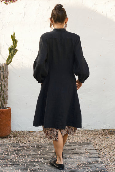 Person wearing a black coat with a pattern skirt   standing against a white wall.