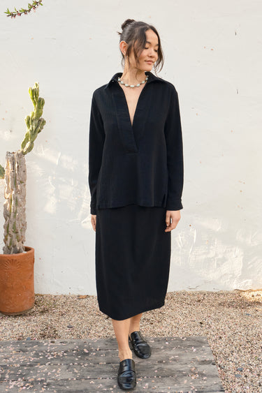Woman wearing a black outfit standing outdoors with a white wall and plant in the background