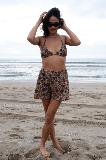 A person wearing sunglasses and a floral patterned woven swimsuit shorts, posing on a beach with the ocean in the background.