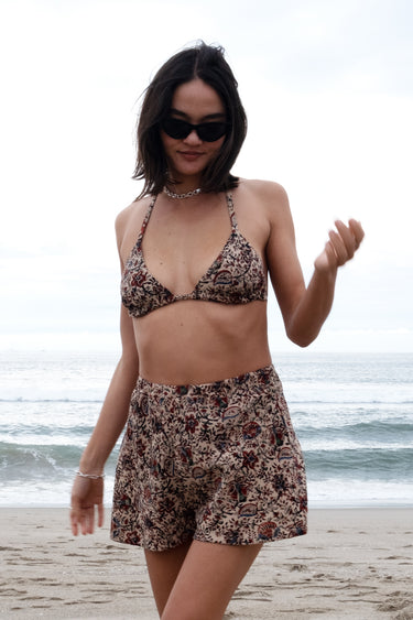 A person wearing sunglasses and a floral patterned woven swimsuit shorts, posing on a beach with the ocean in the background.