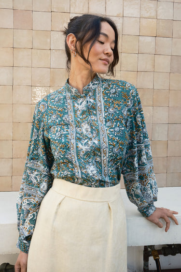 Woman wearing a patterned blouse and cream skirt against a tiled wall.