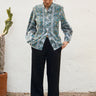 Person wearing a patterned shirt and black pants standing against a white wall with a plant in the background.