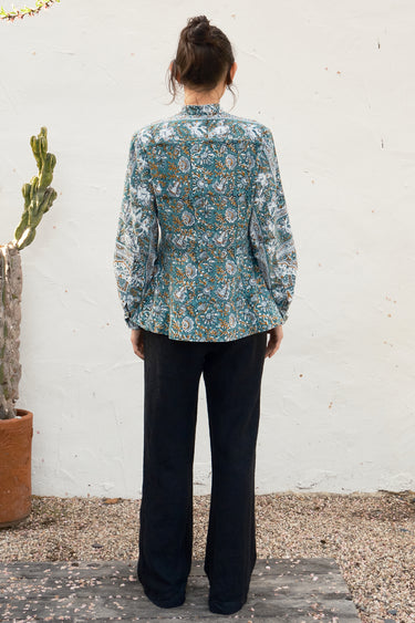 Person wearing a floral blouse and black pants standing against a white wall.