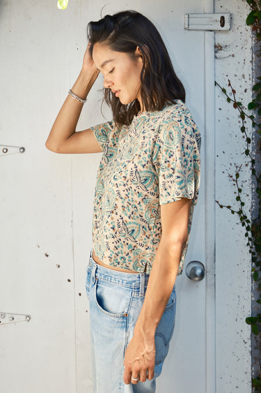 A woman standing with one hand in her hair, wearing a beige and green hand woven and hand block printed Indian tapestry top with short sleeves and a round neckline.