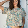 A woman standing with one hand in her hair, wearing a beige and green hand woven and hand block printed Indian tapestry top with short sleeves and a round neckline.