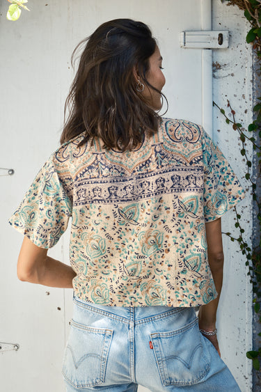 A woman wearing a beige and green hand woven and hand block printed Indian tapestry top with short sleeves and a round neckline. back view