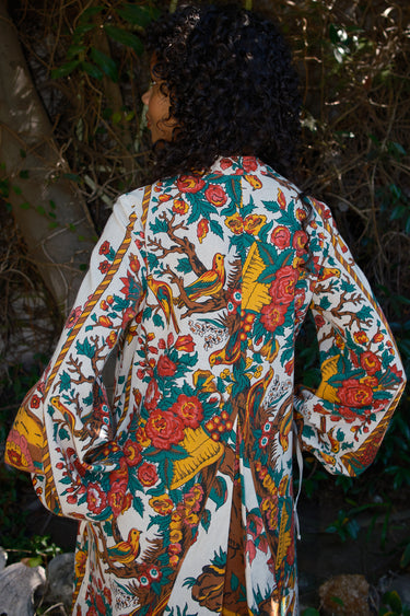 Person wearing a colorful floral and bird patterned caftan in a natural setting.