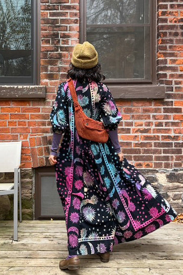 Woman wearing a tapestry maxi caftan. The tapestry is black ground printed with rainbow suns, moons and stars, back view. 