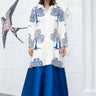 A woman stands against a white wall wearing a long white coat with blue tree-like floral prints and a bright blue caftan paired with black ankle boots. A stylized bird mural is visible on the wall.