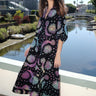 Woman standing on a bridge over a canal wearing a tapestry maxi caftan. The tapestry is black ground printed with rainbow suns, moons and stars.