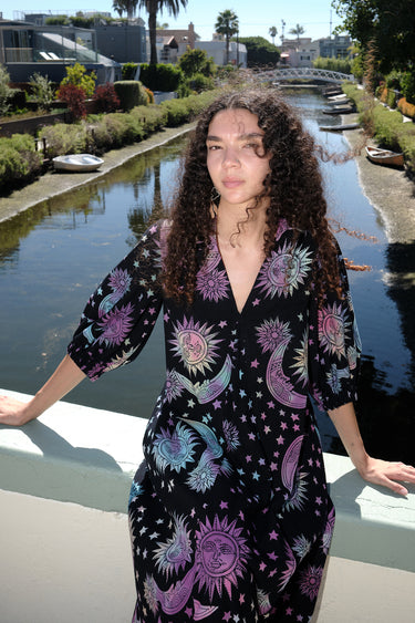 Woman standing on a bridge over a canal wearing a tapestry maxi caftan. The tapestry is black ground printed with rainbow suns, moons and stars.