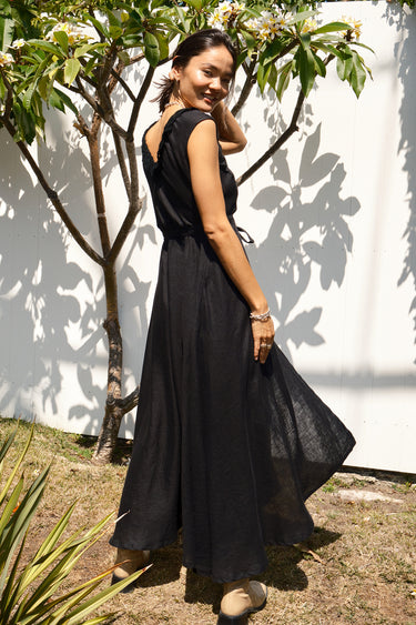A woman standing in a garden wearing a black, wrap-style dress with an elastic neckline and battenberg lace detailing, paired with tan boots. side view 