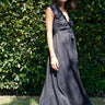 A woman standing in a garden wearing a black, wrap-style dress with an elastic neckline and battenberg lace detailing, paired with tan boots.