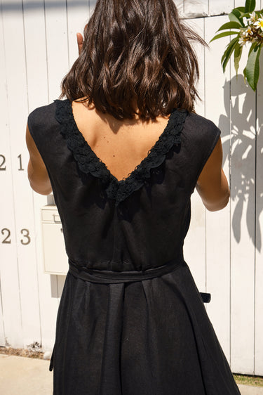 A woman standing in a garden wearing a black, wrap-style dress with an elastic neckline and battenberg lace detailing, back view