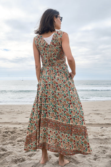Woman standing on the beach wearing a red,  beige and green tapestry maxi wrap dress with a white tank underneath, back view.