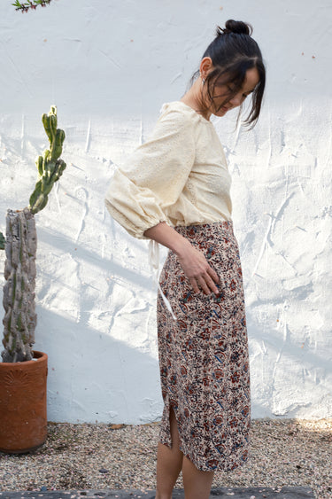 Woman in a floral skirt and beige top standing against a textured white wall with a plant.