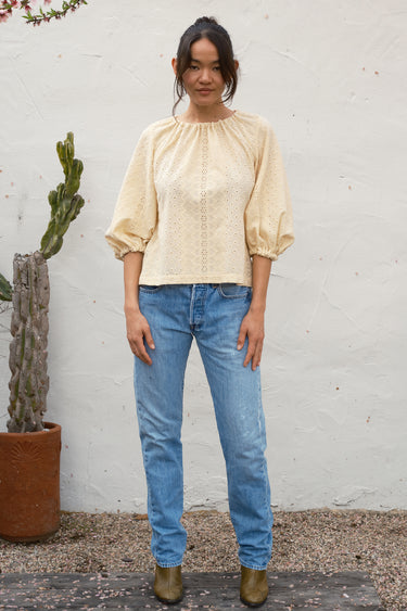 Woman wearing a beige blouse and blue jeans standing against a white wall with a plant in the background.